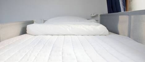 Una cama bien hecha con colchón y almohada blancos, cabecera gris y mesita de noche blanca con una lámpara.