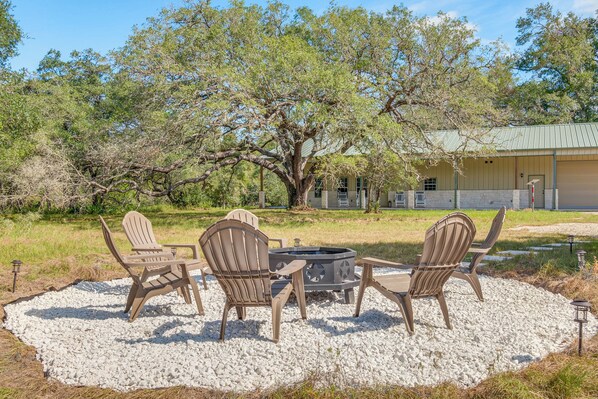 Een terras met Adirondack-stoelen en een vuurkuil, omgeven door een grindgebied met bomen op de achtergrond.