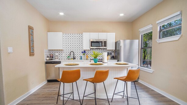 A modern kitchen with white cabinets, a central island with stools, and a view of the outdoors through a window.