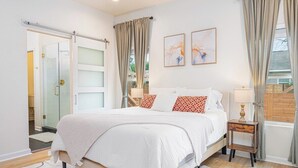 A bedroom with a bed, a nightstand, a window with curtains, and a door leading to a bathroom.