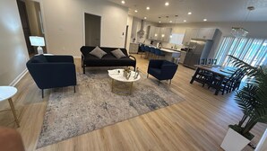 A modern living room with a black sofa, two armchairs, and a coffee table. A kitchen with white cabinets and a dining area with a wooden table and chairs are visible.