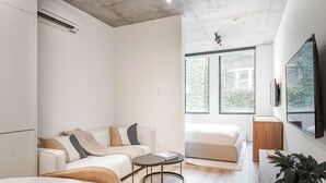 A modern, well-lit room with a sofa, a small round table, a bed, and a large window offering a view of greenery outside. - Modern Studio Haven in Le Mont (Montreal)