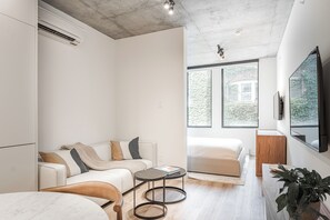 A modern, well-lit room with a sofa, a small round table, a bed, and a large window offering a view of greenery outside. - Experience Montreal Living at Le Mont (Montreal)