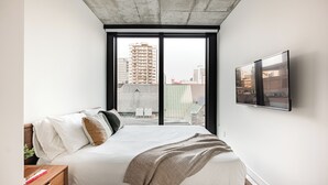 Un dormitorio moderno con una cama grande, una mesita de noche de madera, un televisor montado en la pared y vistas de la ciudad a través de amplias ventanas.