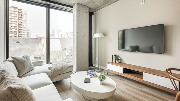 Una sala de estar moderna con un ventanal grande, un televisor de pantalla plana, un sofá blanco, una mesa de centro y una mesita de noche de madera.
