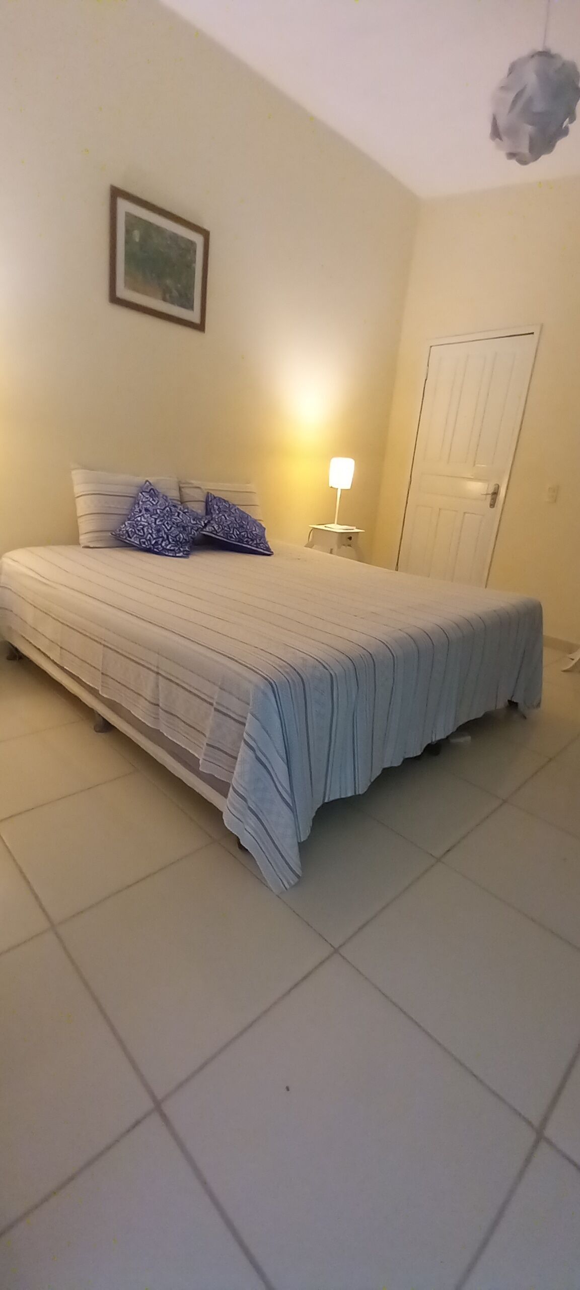 Un dormitorio con cama, mesita de noche, lámpara, una puerta y un cuadro en la pared.