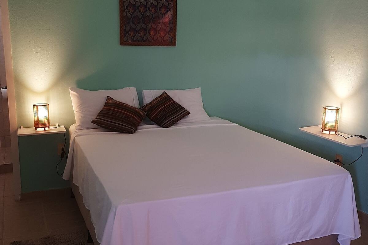 A neatly made bed with white bedding and two decorative pillows, flanked by two bedside tables with lamps, in a room with teal walls and a single artwork above the bed.