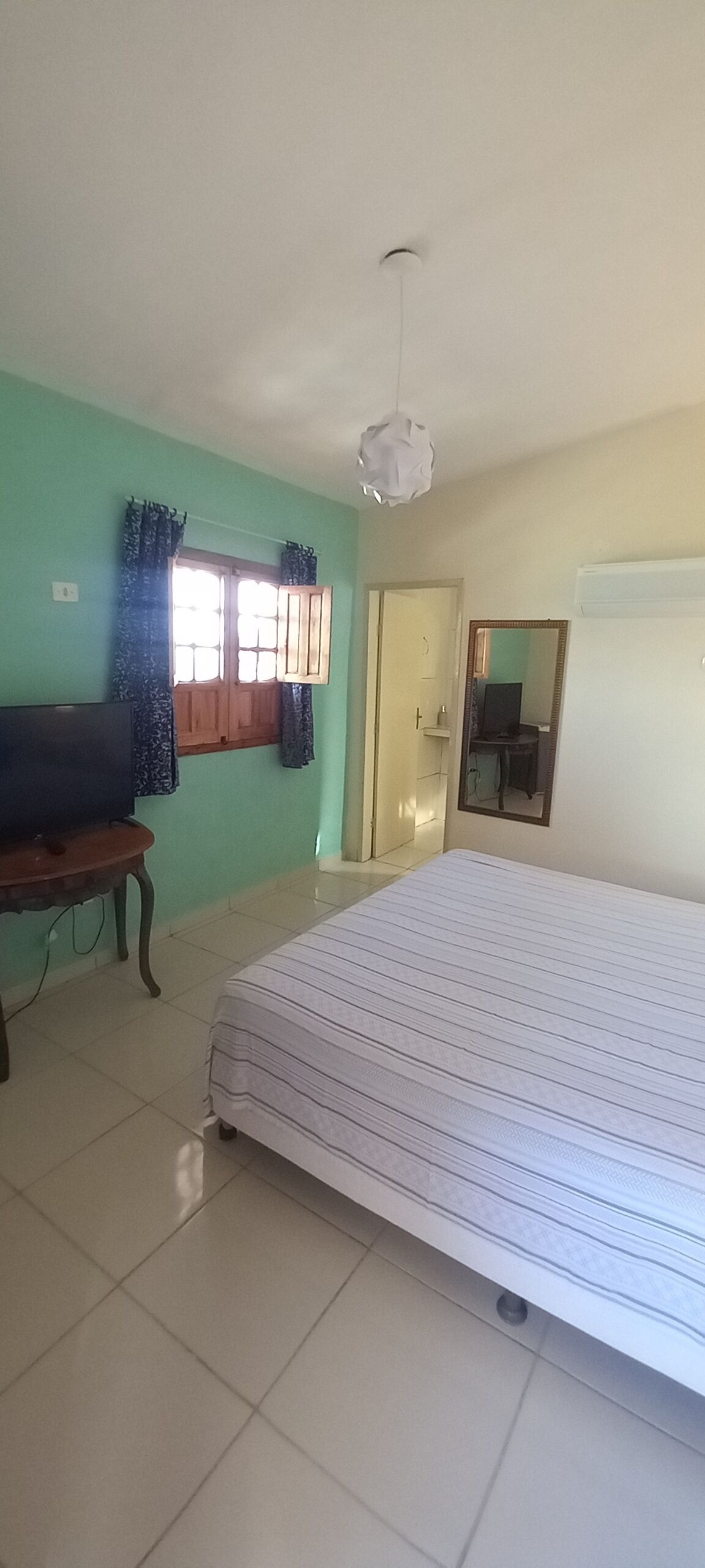 A bedroom with a bed, a television on a wooden table, a window with curtains, and a door.