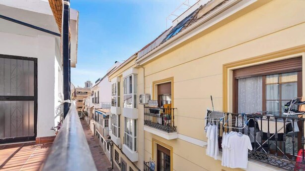 A narrow urban street with multi-story buildings, balconies with clothes drying, and a clear sky.