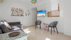 A modern living room with a grey sofa, patterned coffee table, and two black chairs. A flat-screen TV is mounted on the wall, and a wooden shelf with a giraffe figurine is visible.
