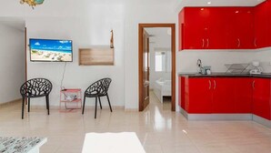 A modern living room with a TV, two chairs, a small table, and a red kitchen area.