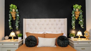 Un dormitorio con cabecera capitonada, dos mesitas de noche con lámparas, un reloj y plantas decorativas.