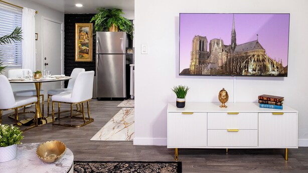 Una cocina moderna con un refrigerador de acero inoxidable, un área de comedor con sillas blancas y una mesa dorada, y un armario blanco con tiradores dorados.