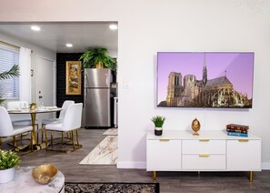Una cocina moderna con un refrigerador de acero inoxidable, un área de comedor con sillas blancas y una mesa dorada, y un armario blanco con tiradores dorados.
