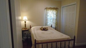A bedroom with a bed, a nightstand, a lamp, a window with curtains, and a door. - Cedar Grove Cottage Conveniently Located to all attractions. (New Bern)