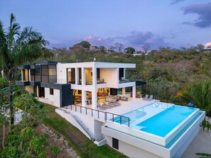 Una casa moderna de dos pisos con una amplia piscina, rodeada de exuberante vegetación y colinas.
