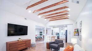 Ein modernes Wohnzimmer mit Flachbildfernseher, Holzbalken an der Decke, einem weißen Sofa, einem grauen Sessel und einem runden Hocker.