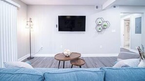A modern living room with a flat-screen TV mounted on the wall, a small wooden table with a decorative bowl, and a blue sofa.