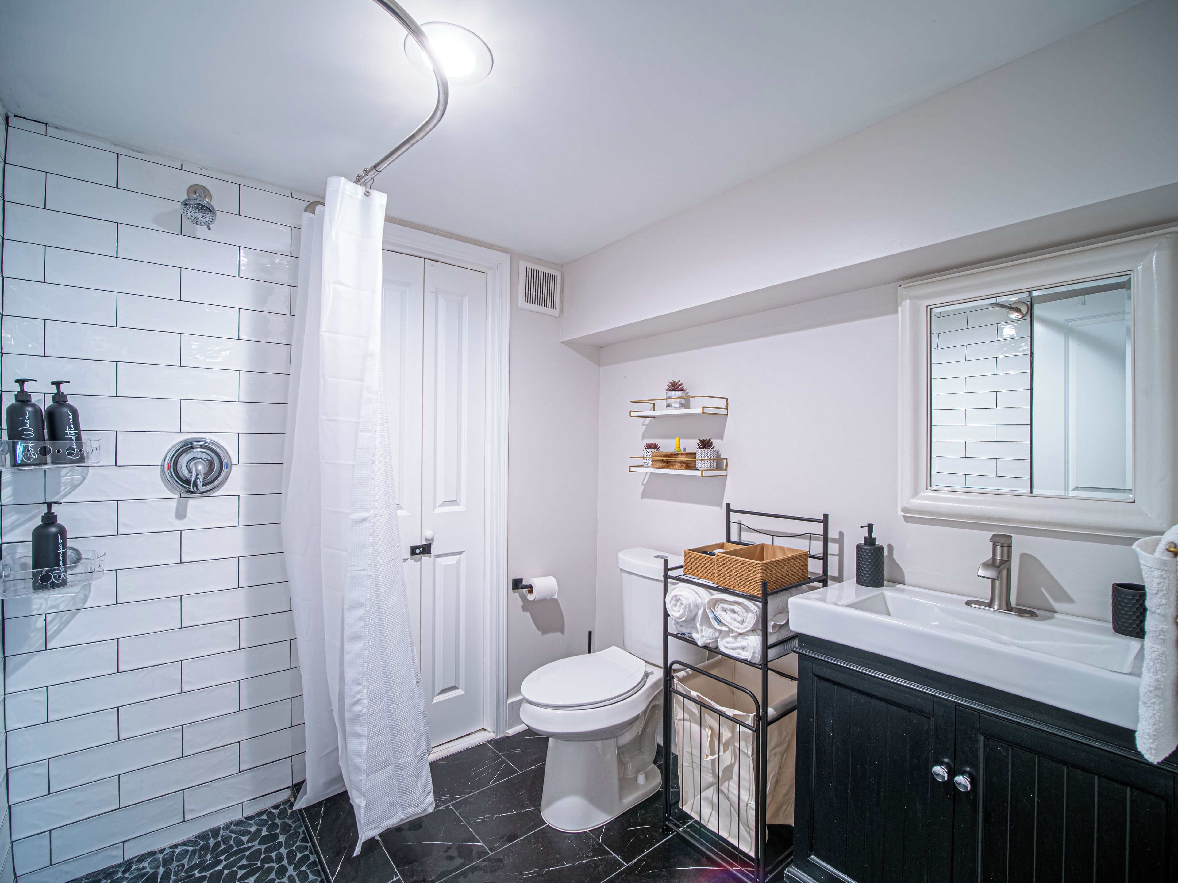 Un baño con paredes de azulejos blancos, una ducha con cortina, un inodoro, un lavabo con espejo y repisas con artículos de tocador.