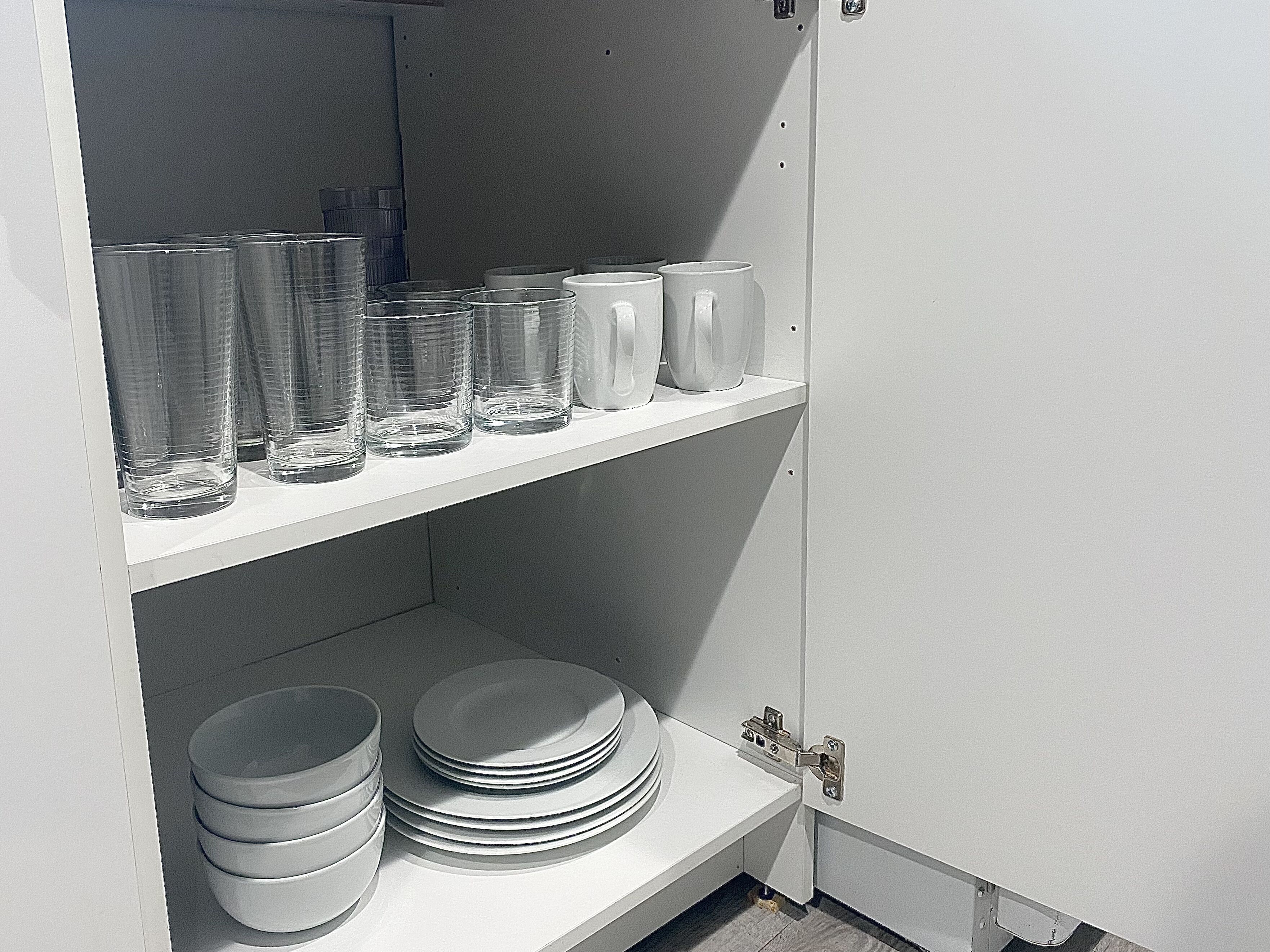 A kitchen with white cabinets, a white countertop, and shelves holding clear glasses, white mugs, and stacked white plates.