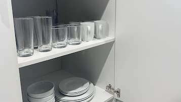 A kitchen with white cabinets, a white countertop, and shelves holding clear glasses, white mugs, and stacked white plates.