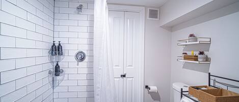 Une salle de bain avec des murs carrelés de carreaux blancs, une douche, des toilettes et des étagères contenant des articles de toilette.