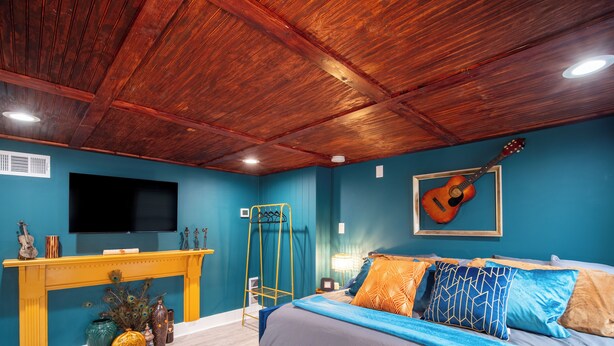 Een slaapkamer met een houten plafond, een bed met blauwe en oranje beddengoed, een televisie aan de muur en een gitaar.