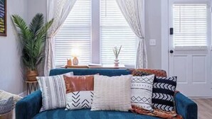 A living room with a blue sofa, patterned cushions, a potted plant, and a window with white curtains. - Lumiére of Marietta Square (Marietta)