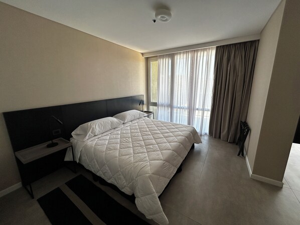 Une chambre d’hôtel moderne avec un grand lit, une table de chevet, une lampe et une vue sur l’extérieur à travers des rideaux transparents.