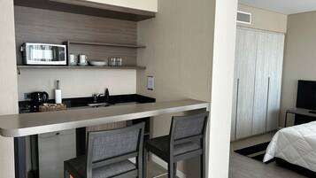 Une petite cuisine équipée avec un micro-ondes, un évier et des étagères de rangement. Il y a deux chaises et un lit avec une couette blanche.