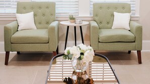 Two green armchairs with white cushions, a small round table with a vase of flowers, and a window with blinds in the background. - The Grand Slam - The Battery ATL (Smyrna)
