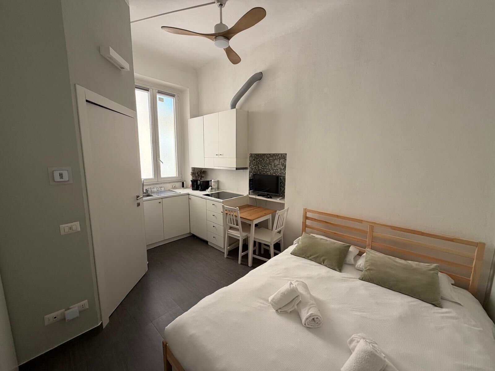 A compact hotel room with a bed, a desk, a chair, a small kitchenette, and a ceiling fan.