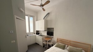 Kompakti hotellihuone, jossa on sänky, työpöytä, tuoli, pieni keittokomero ja kattotuuletin.