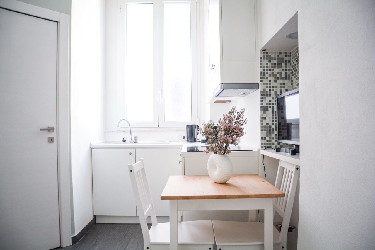 Eine kleine, helle Küche mit weißen Schränken, einem Fenster und einem Holztisch mit Blumen in einer Vase.