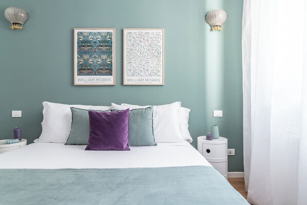 Un lit bien fait, avec une literie blanche et turquoise, deux œuvres d’art encadrées au-dessus, et une table de chevet avec une lampe et un vase.