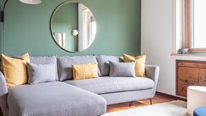A grey sectional sofa with yellow cushions, a round mirror, and a black floor lamp against a green wall. - [Vista Duomo] Trilocale vintage vicino a Centrale (Milan)