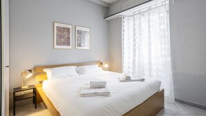 Een keurig opgemaakt bed met gevouwen handdoeken, een nachtkastje met een lamp, en twee ingelijste kunstwerken aan de muur.