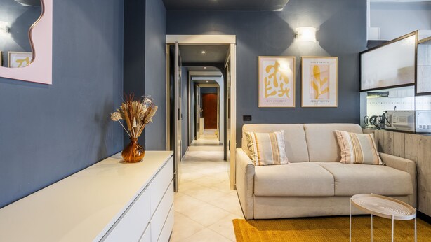 Een moderne woonkamer met een bank, een bijzettafeltje en een vaas met gedroogde bloemen. Een gang leidt naar een andere kamer.