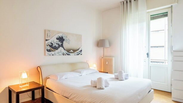 A neatly made bed with white linens and towels, a bedside table with a lamp, a wall-mounted artwork, and a view of the outside through a door. - [Niguarda] Bright and comfortable Flat (Milan)