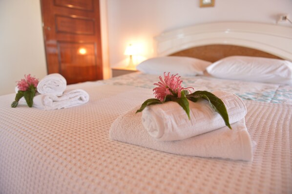 A neatly made bed with rolled white towels and pink flowers, a wooden door, and a nightstand with a lamp.