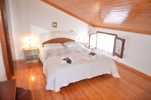 Un dormitorio con techo de madera, una cama con cubrecama blanco, una mesita de noche con lámpara y una ventana con vistas.