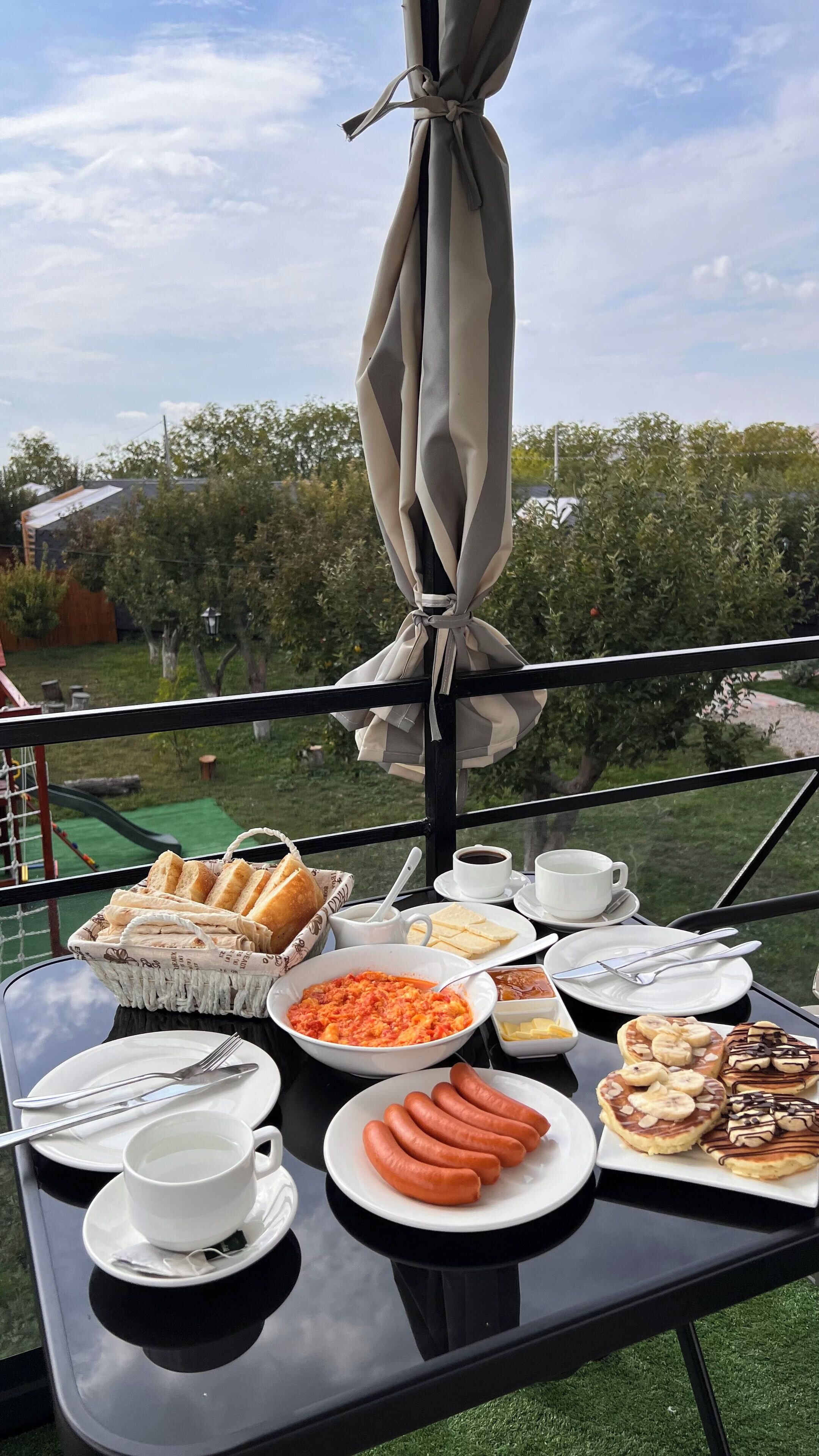 A table set with breakfast items including bread, sausages, and pastries, with a view of trees and a clear sky.
