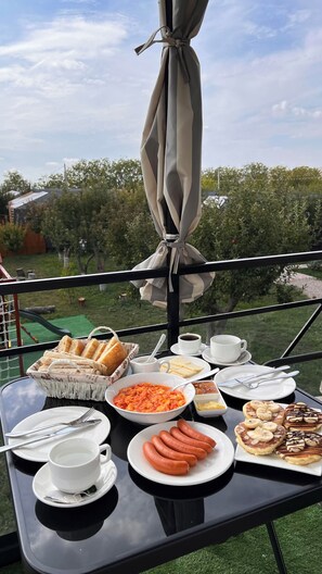 A table set with breakfast items including bread, sausages, and pastries, with a view of trees and a clear sky. - Hidden Garden Resort (Garni)