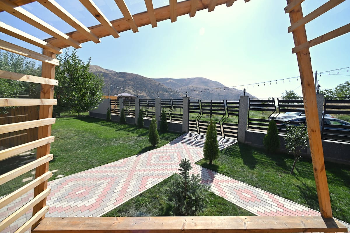 A wooden pergola structure with a paved pathway leading to a fenced area with a mountain backdrop.