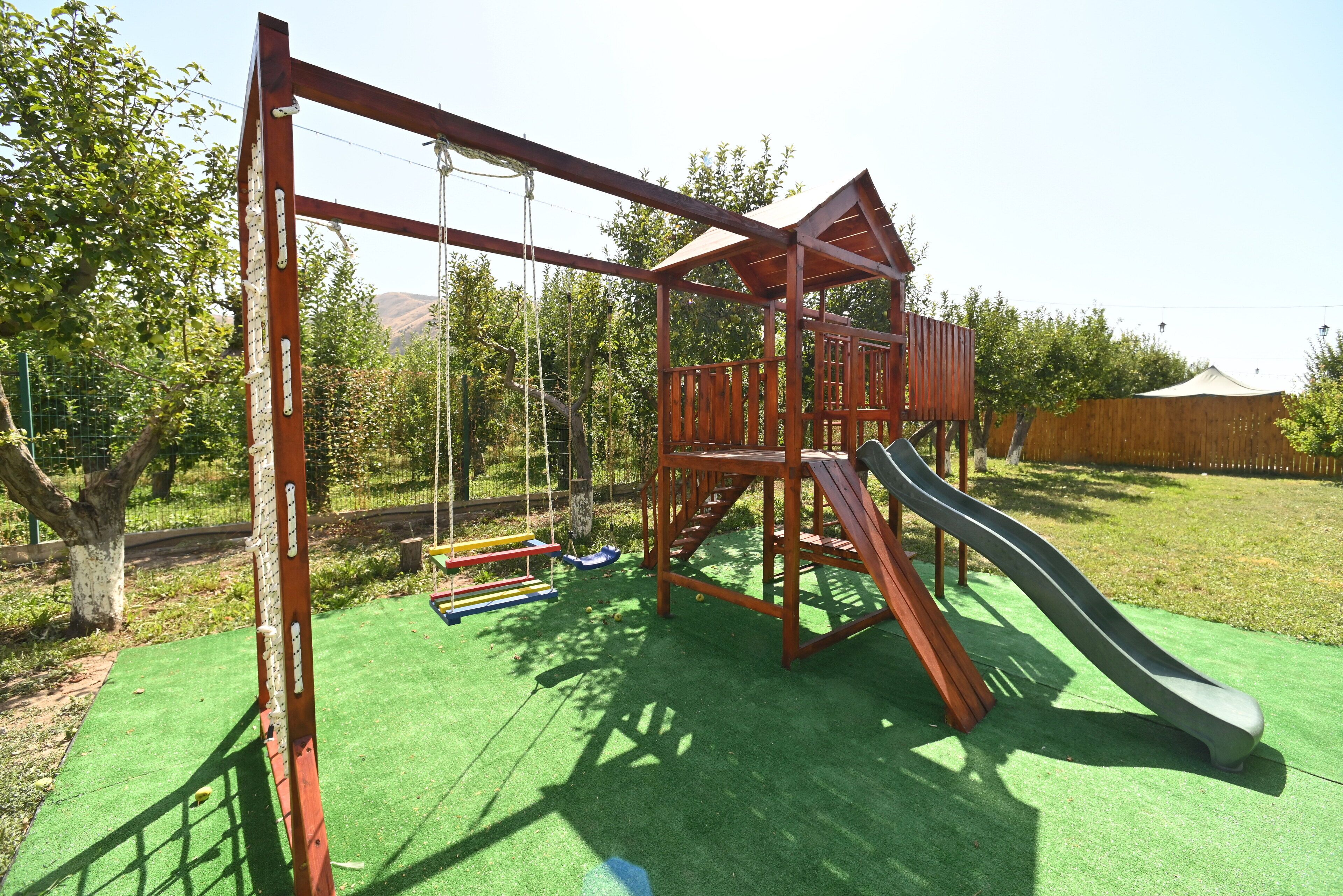 A wooden play structure with a slide and swings on green artificial turf.