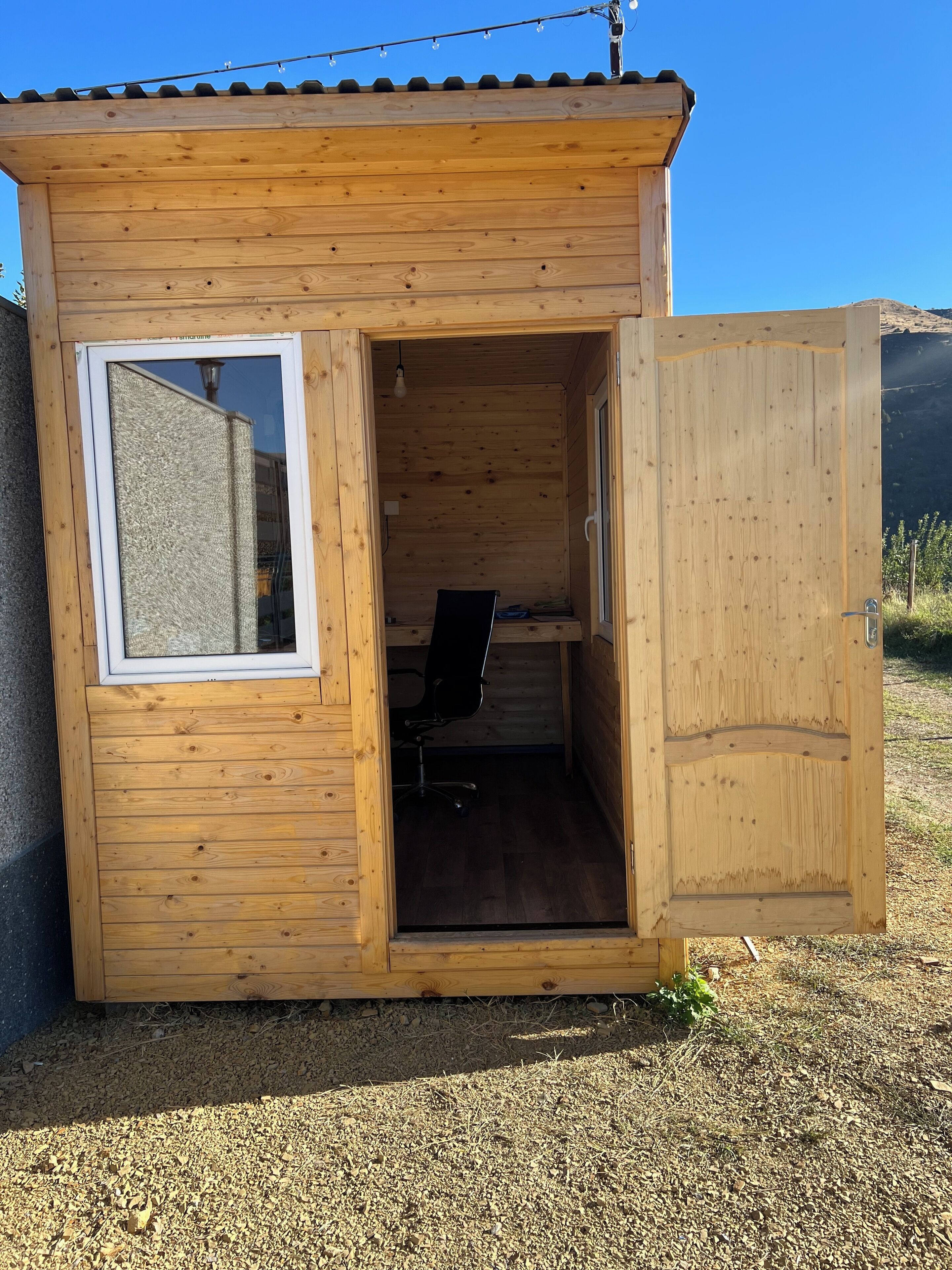 Una piccola baita di legno con una sola porta aperta, da cui si intravede una scrivania e una sedia all'interno.