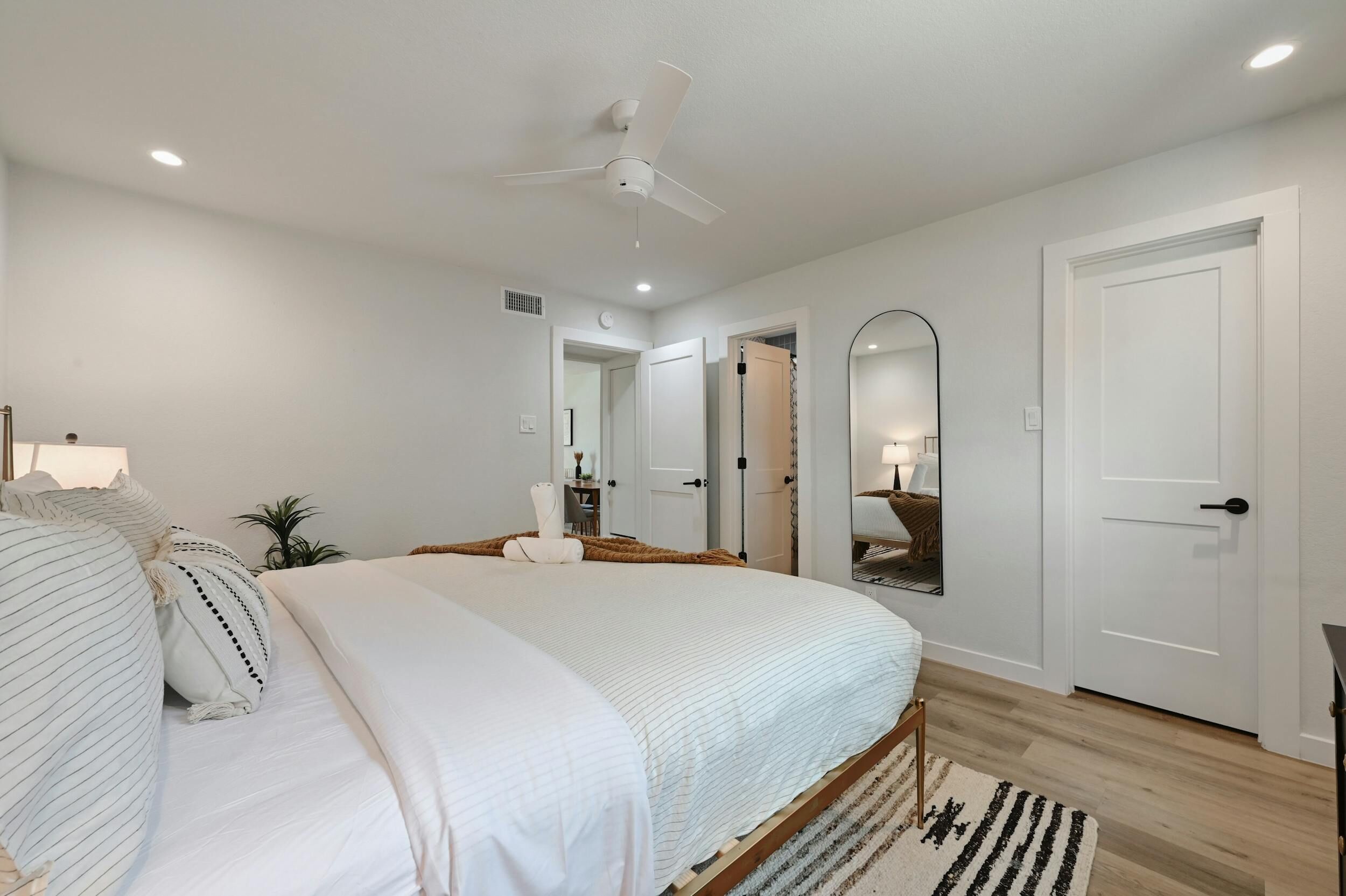 A bedroom with a large bed, a ceiling fan, a mirror, and a door.