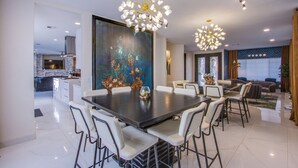 A modern dining area with a large table and chairs, a painting on the wall, and a kitchen in the background.