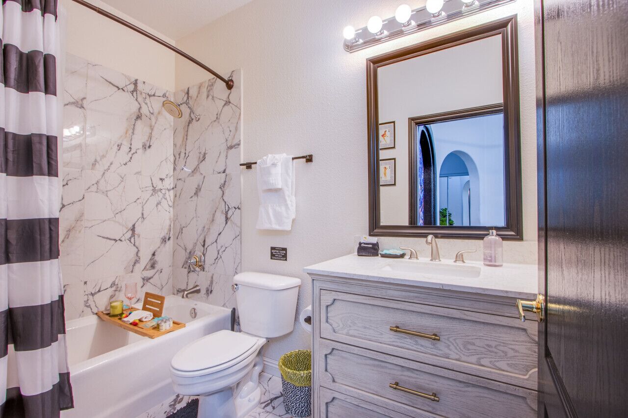 A bathroom with a bathtub, toilet, vanity with drawers, a mirror with lights, and a marble wall.
