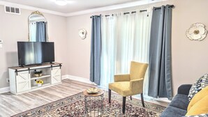 Una sala de estar con televisor sobre un mueble de madera, un sillón amarillo, una alfombra estampada y un ventanal con cortinas.